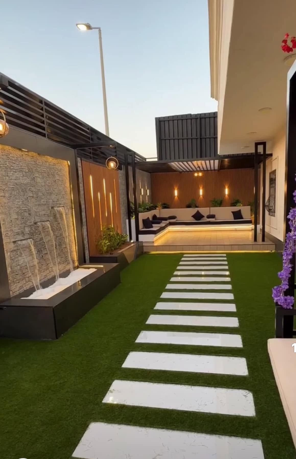 Evening outdoor lounge with a lit waterfall wall, pergola seating and a turf walkway