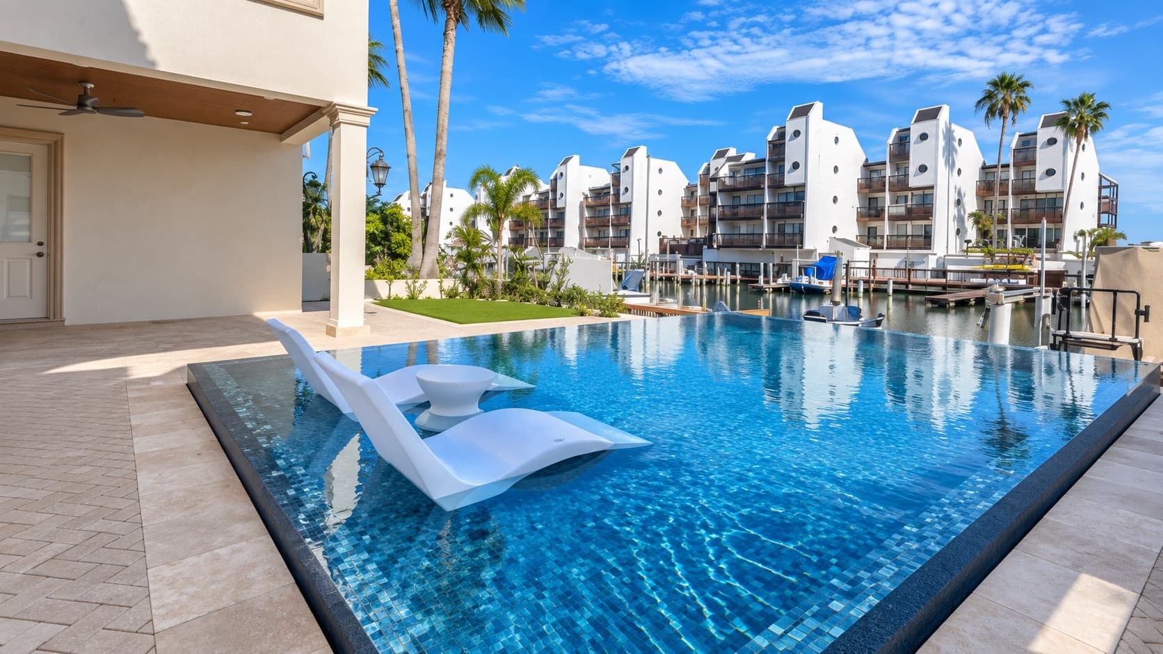 Glass-tile infinity pool with in-water loungers and travertine deck overlooking a marina